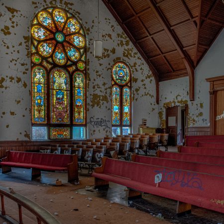 Abandoned church with a wall of stained glass, red pews, wooden seating and a cathedral ceiling.
