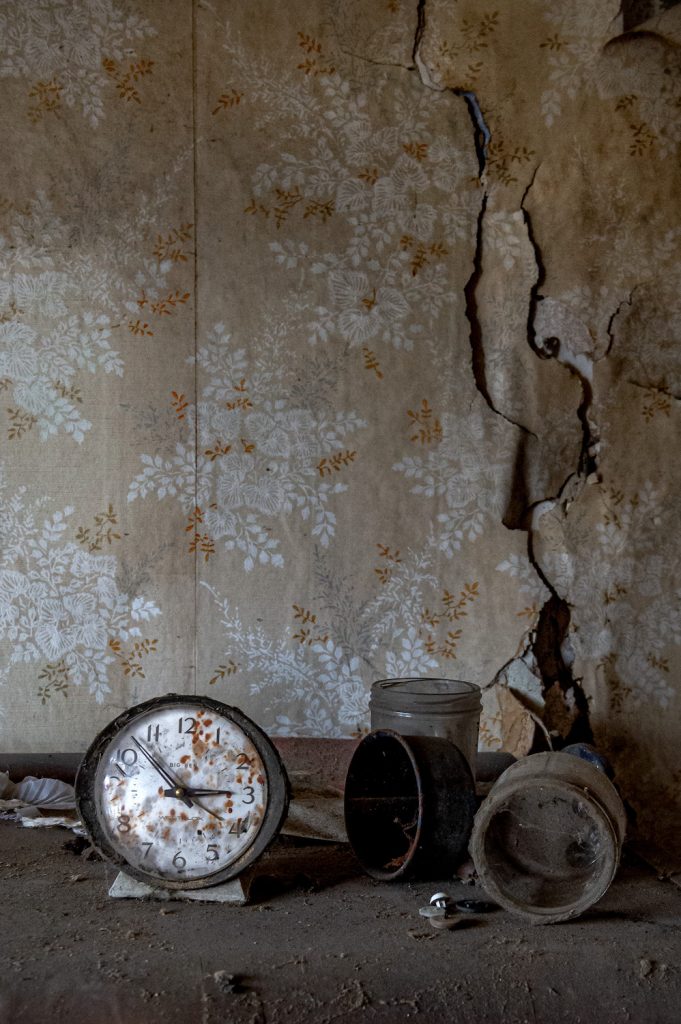 Abandoned dining room with clock and glassware collecting dust on the sideboard with cracked wallpaper as a background.