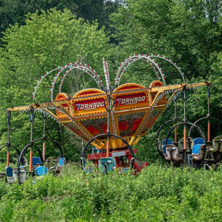 A Tornado tilt-a-whirl amusement park ride sits abandoned in a field of weeds and trees.